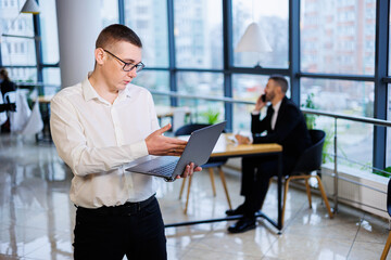 A young businessman man in glasses and a white shirt with a laptop in his hands stands in the office and works