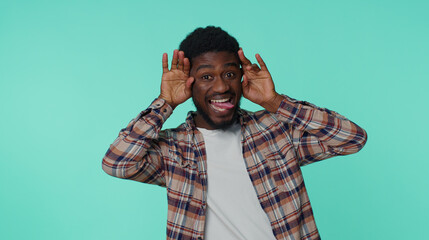 Funny happy sincere adult man 20 years old in shirt making playful silly facial expressions and grimacing, fooling around showing tongue. Young african american guy alone on blue wall background