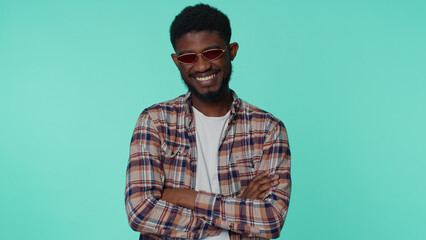 Fototapeta premium Portrait of seductive cheerful casual african american man wearing red sunglasses, charming smile. People emotions concept. Young lovely adult guy posing isolated alone on blue studio wall background