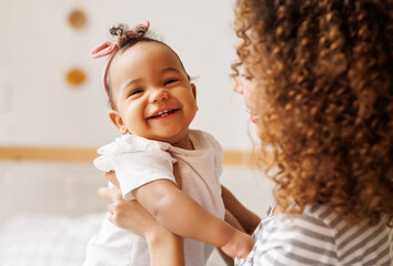 Happy, cheerful ethnic mom holds a laughing baby daughter in her arms. - 491517586