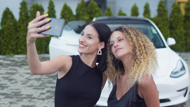 Two beautiiful girls in black outfut smiling and posing for a selfy photo in front of fancy sports car..