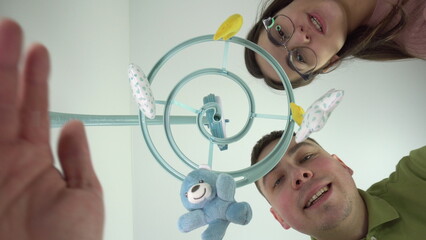 Young parents look at a newborn baby lying in a cradle with a baby mobile with toys. First-person...