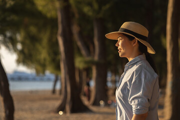 Young asian woman with straw hat standing alone on beach looking at sunset seashore. Chilling in holiday weekend summertime. Traveler Female walking around the beach with the sunlight