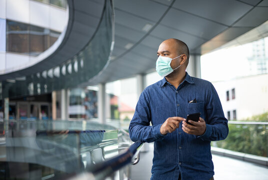 Young indian using smartphone standing in front of office building. Asian man wearing protective mask during pandemic.