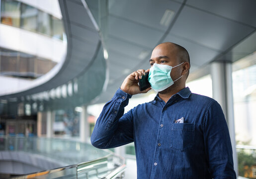 Young indian using smartphone standing in front of office building. Asian man wearing protective mask during pandemic.