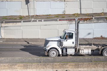 Day cab white big rig semi truck with chrome accessories and high exhaust pipes transporting cargo in semi trailer running on the highway road with concrete wall on the side
