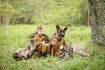 a boy in a military uniform in a clearing, sitting by a campfire with a German shepherd.Two friends defend the motherland