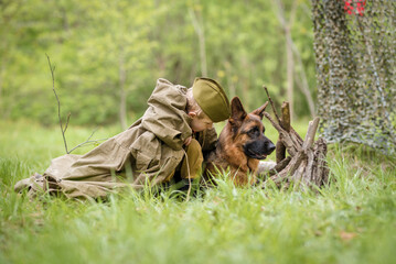 a boy in a military uniform in a clearing, sitting by a campfire with a German shepherd.Two friends defend the motherland