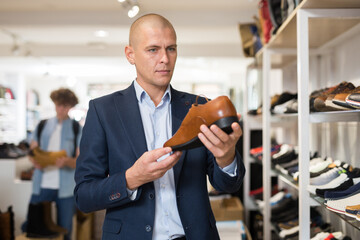 Portrait of focused man choosing formal shoes in store, looking with interest at stylish brown leather dress shoe with lacing