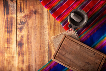 Mexican food cooking background with empty cutting board, colorful traditional fabric and molcajete on rustic wooden table. Top view with copy space.