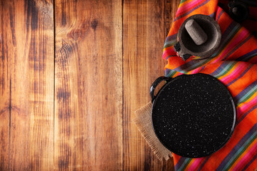 Mexican kitchen utensils, colorful traditional fabric, comal de peltre and molcajete on wooden rustic table. Top view with copy space