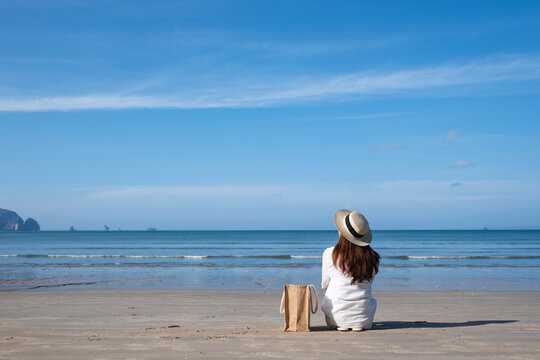 Rear view image of a woman with hat and bag sitting on the beach with blue sky background