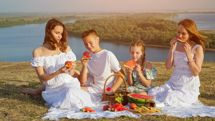 Mother and happy children have festive picnic with tasty food sitting on white tablecloth on green brae against tranquil wide river on sunny summer day, sunlight