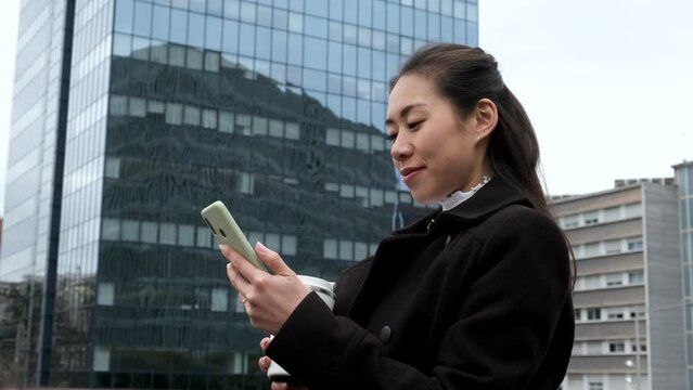 Low angle side view of glad Chinese female manager with coffee to go. She is smiling and browsing mobile phone while standing against glass skyscraper on city street
