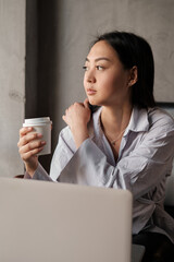 A businesslike Asian woman, working on a laptop and holding a paper cup with tea or coffee in her hand.