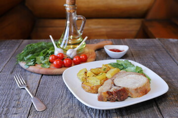 On a white ceramic plate are a big chunk of baked pork with rustic fried potatoes on the background of a wooden cutting board with tomatoes, herbs and a jug of oil. 