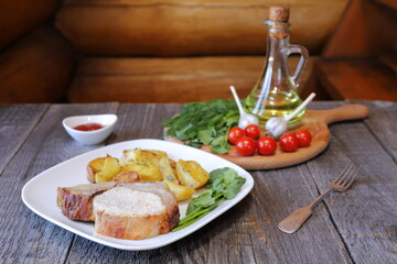 On a white ceramic plate are a big chunk of baked pork with rustic fried potatoes on the background of a wooden cutting board with tomatoes, herbs and a jug of oil. 