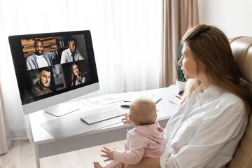 Lovely mother in maternity leave engaged in remote work or study at home on PC while infant baby girl hold documents and looking at screen. Modern day mother of little kid sit at desk use gadgets