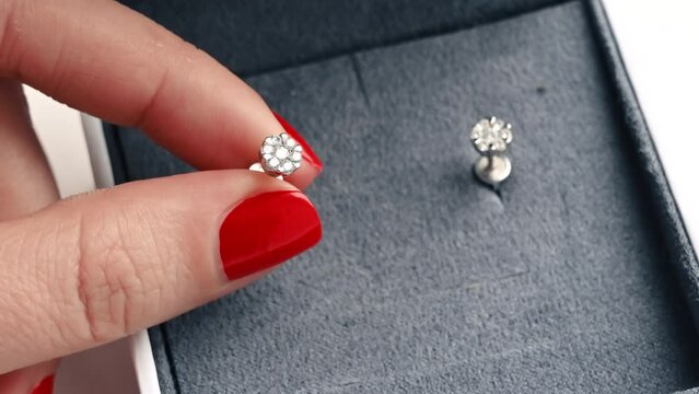 Close view of a woman holding earring with diamonds. A box with another earring, hands with red nails
