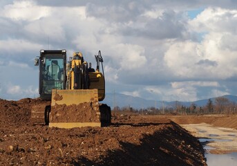 Front view of an excavator, with the lighted empty cabin, between a cloudy sky and the dirt soil of...