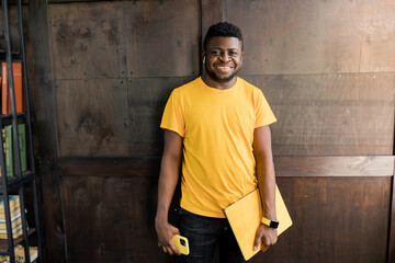 Portrait of an attractive smiling confident casual young man looking at camera, college student or freelancer, standing over background, carrying yellow laptop computer in hands