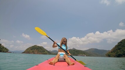 Pretty sexy young blonde woman in stylish swimsuit and sunglasses enjoys trip canoe on sea against palm islands. Traveling to tropical countries. Positive sport girl hand padding on kayak front view.