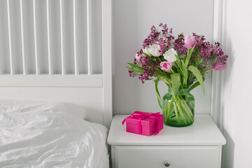 Pink box present and vase with spring flowers on bedtable near bed. Scandinavian Interior.