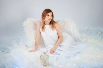 An attractive girl in a white tunic with large white wings behind her back poses while sitting in white luminous clouds.
