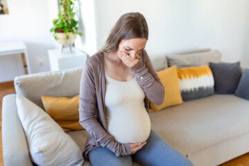 Pregnant woman suffering during her pregnancy, with back pain and headaches. Pregnant woman sitting on the sofa holding her belly with worried face expression. - obrazy, fototapety, plakaty