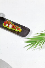 Modern food - tuna tartare on sliced avocado in black plate. Avocado starter with tuna and radish in asian style. Contemporary asian dish. Healthy food with avo and marinated fish on white background.