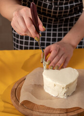 Woman's hand holds palette knife over heart-shaped bento cake. The cake stands on parchment and wooden board. Close-up. Selective focus.