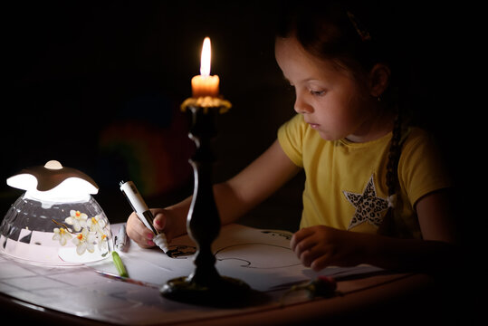 Little girl draws fantasy characters near candle in a dark room. Child doing favorite thing during a power outage