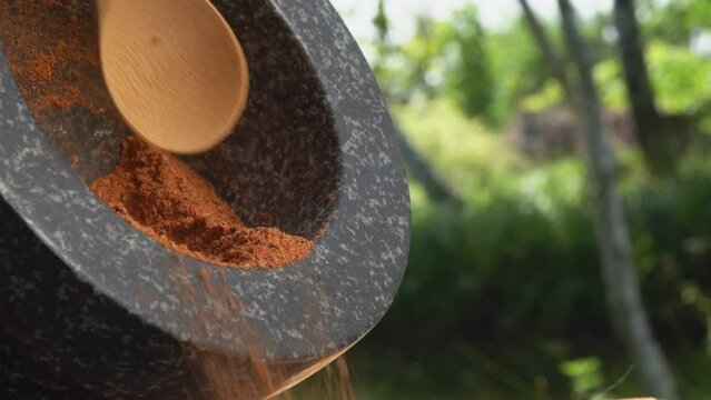 Bamboo spoon is taking a red mix of spices and peppers from a grey stone mortar