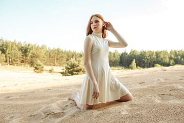 Fototapeta na wymiar Stylish woman with long red hair and in summer dress standing on knees on sand in nature and looking away 