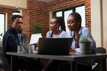 Company office employees cooperating regarding startup project marketing expenses while in modern office interior. African american business people discussing financial strategy while in workspace.