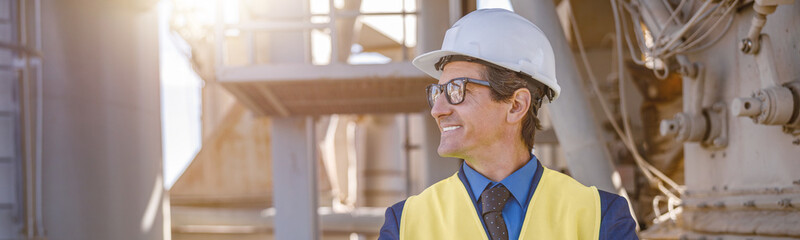 Joyful matured man wearing safety helmet and work vest while keeping arms crossed and smiling