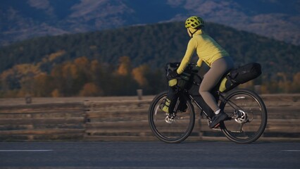 Fototapeta premium The woman travel on mixed terrain cycle touring with bikepacking. The traveler journey with bicycle bags. Sport tourism bikepacking, bike, sportswear in green black colors. Mountain snow capped.