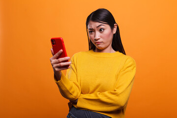 Doubtful uncertain beautiful woman wearing yellow sweater while using smartphone device on orange background. Confused clueless adult person using mobile cellphone to understand un clear instructions. - obrazy, fototapety, plakaty