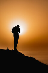 Silhouette of a man taking pictures from a cliff watching the ocean with the sunset in the background.