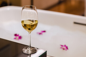 A glass of white wine puts on table beside bathtub that have white bubble and orchid flowers....