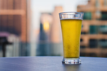 A glass of beer puts on table at rooftop bar with blurred city background.