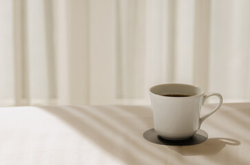 A cup of black coffee puts on white bed sheet with shadow of curtain in the morning. Relaxing at home concept.
