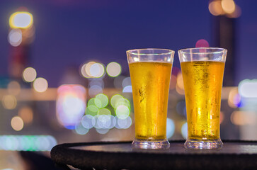 Two glasses of beer put on table at rooftop bar with colorful city bokeh light background.