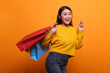 Overjoyed excited beautiful asian woman carrying shopping bags from outlet store while wearing yellow sweater. Happy positive fashionable shopper smiling heartily while on orange background.