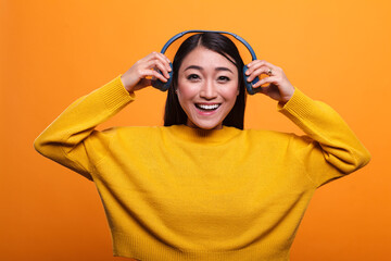 Beautiful smiling heartily woman taking off headphones when she meets her friend. Cute joyful positive asian person stops listening to music to talk with mate while on orange background.
