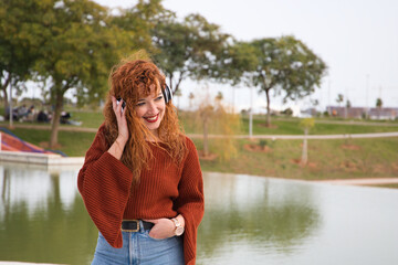 Young and beautiful red-haired woman listening to music with headphones in a park where there is a large lake. The woman is happy and dancing. Concept expressions and happiness, music and dancing.