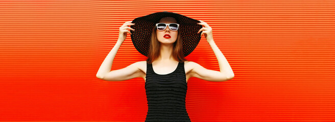 Portrait of beautiful young woman wearing a black summer hat and dress on red background