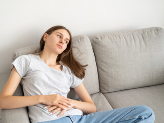 Female disease. Suffering woman. Home recovery. Tired pretty lady feeling pain holding hands on belly sitting sofa in light room interior.