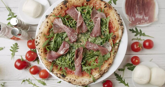 Delicious Pizza with prosciutto parma ham, arugula salad rocket on white background with ingredients. Top view. Italian cuisine. Prosciutto pizza with strachatella cheese, tomatoes, arugula leaves.