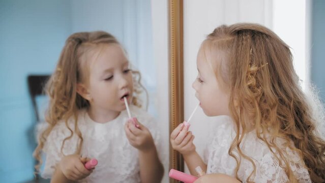 child paints lips. portrait. little cute curly blonde girl paints her lips with lipstick, in front of the mirror.